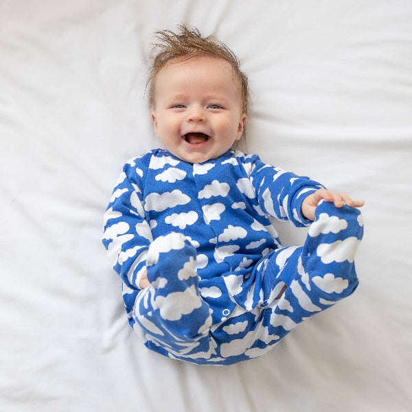 Baby wearing a blue onesie with white cloud patterns, lying on a white surface.