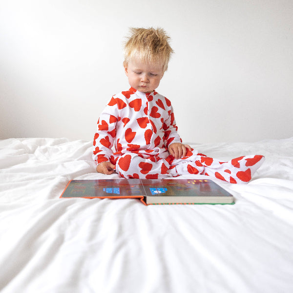 Child wearing a red and white heart-patterned onesie sitting on a bed with a book in front of them.