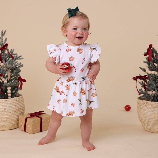 Baby in a white dress with gingerbread pattern standing between two small Christmas trees on a beige background.
