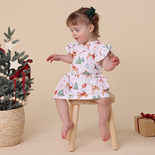 Child in a festive dress standing on a stool next to a decorated Christmas tree and gift box on a beige background