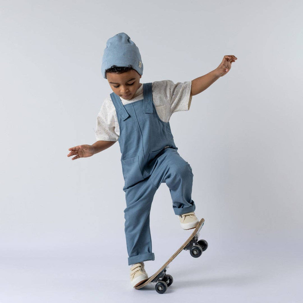 Child wearing blue overalls and a matching beanie skateboarding on a white background