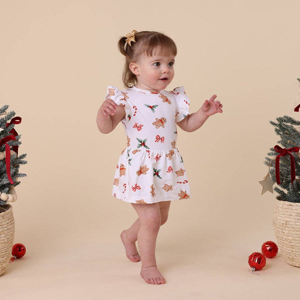 Child wearing a Christmas-themed dress with gingerbread men and candy cane patterns, standing against a beige background.