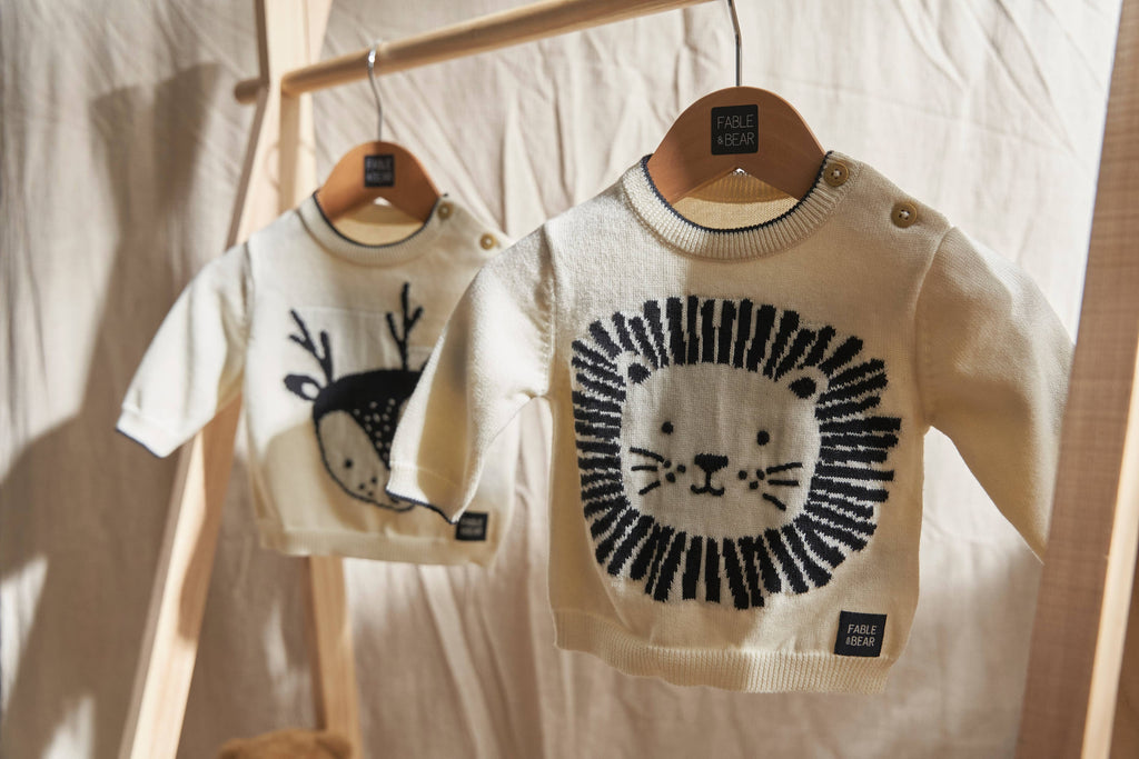 Two children's jumpers/sweaters with animal designs on hangers against a neutral background