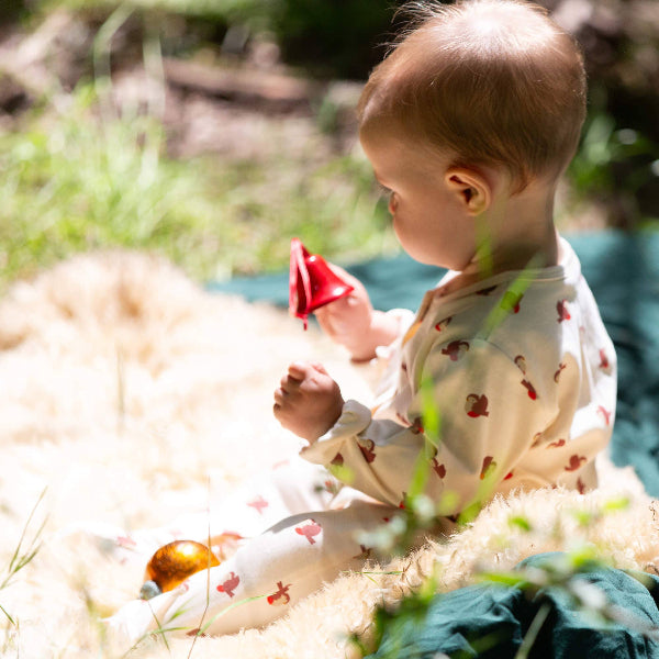 Baby sitting on a blanket outdoors holding a red toy, surrounded by grass and plants.