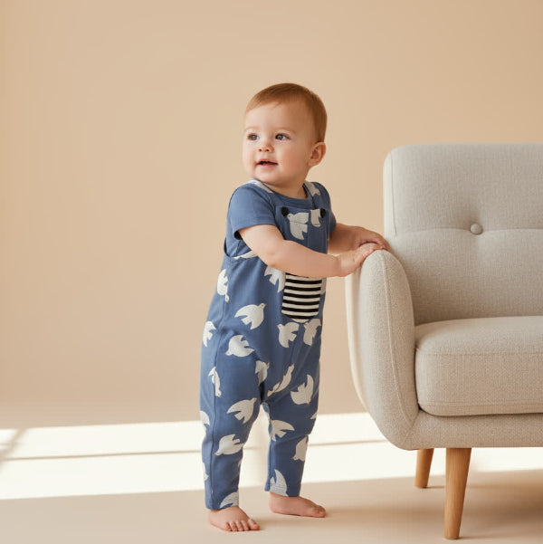 Baby in a blue onesie with white patterns standing next to a light gray chair in a room with beige walls.