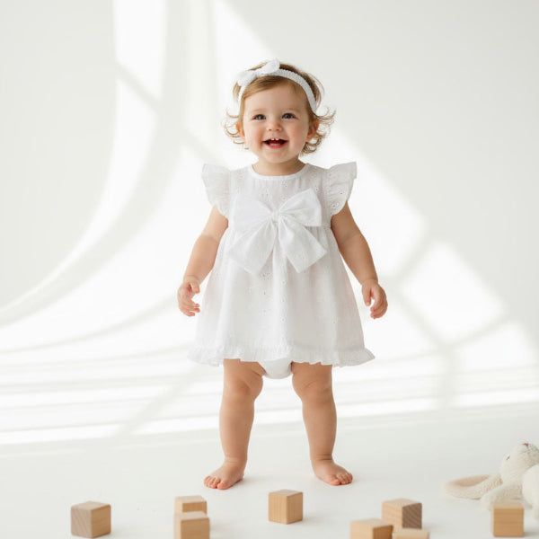 Child in a white dress standing on a white floor with wooden blocks and a stuffed animal.