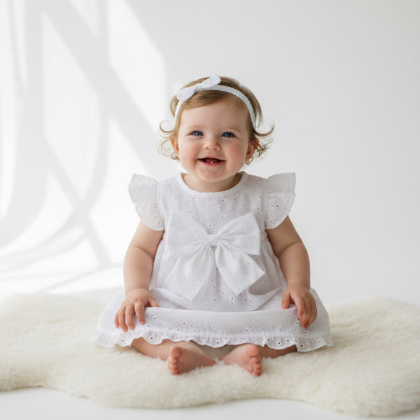 Baby in a white dress with a bow sitting on a white rug against a white background