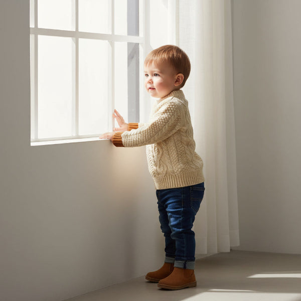 Child in a beige sweater standing by a window in a bright room