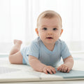 Baby in a light blue onesie lying on a white mat with a white curtain background