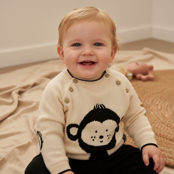 Child wearing a cream sweater with a monkey design, sitting on a beige blanket.