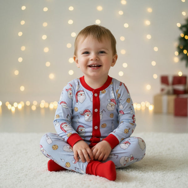 Child wearing Christmas-themed pajamas sitting on a carpet with blurred lights and decorations in the background