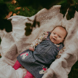Baby girl lying on a cream blanket outdoors in grass, wearing a denim jumper with floral sleeves and bright pink tights.
