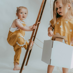 Two young children in mustard outfits engage in pretend play with a cardboard box and a wooden ladder prop in a bright studio.