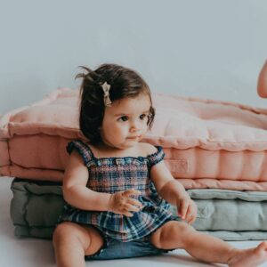Baby girl with a bow in her hair sits on the floor in front of stacked pink and gray blankets, wearing a blue plaid dress.